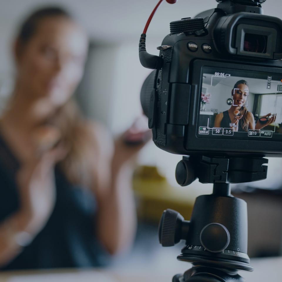 A digital camera on a tripod captures a woman applying makeup. The camera's screen shows a clear view of the woman, who is holding a makeup brush and possibly a compact mirror, while the background is blurred.