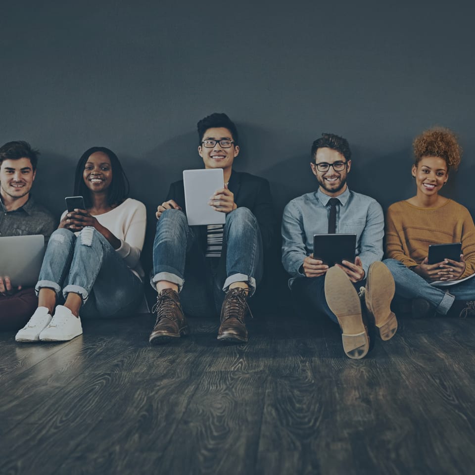 A diverse group of five young adults sitting on the floor against a dark wall, using various electronic devices. They are smiling and appear engaged, holding items like laptops, tablets, and smartphones. The floor is wooden.