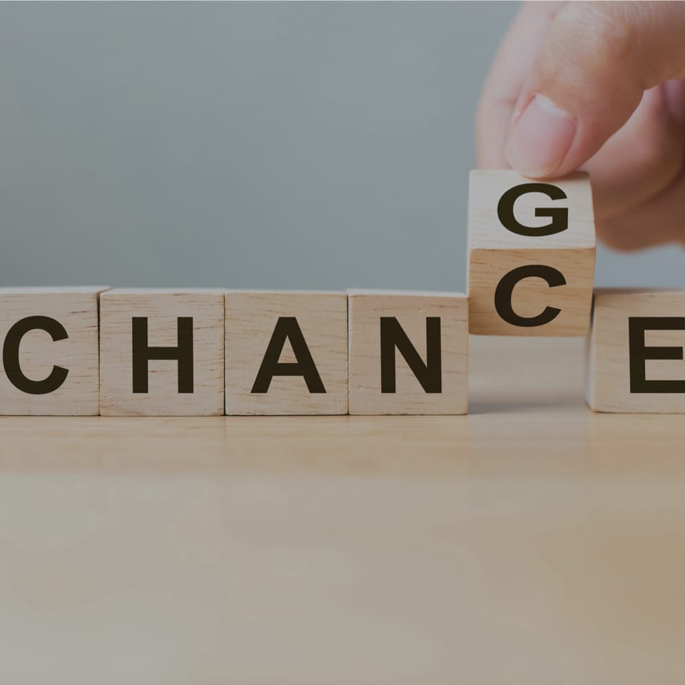 Close-up of wooden blocks on a table spelling out "CHANGE", with a person's hand rotating the last block to reveal a "G", forming the word "CHANCE". The background is plain and grey.