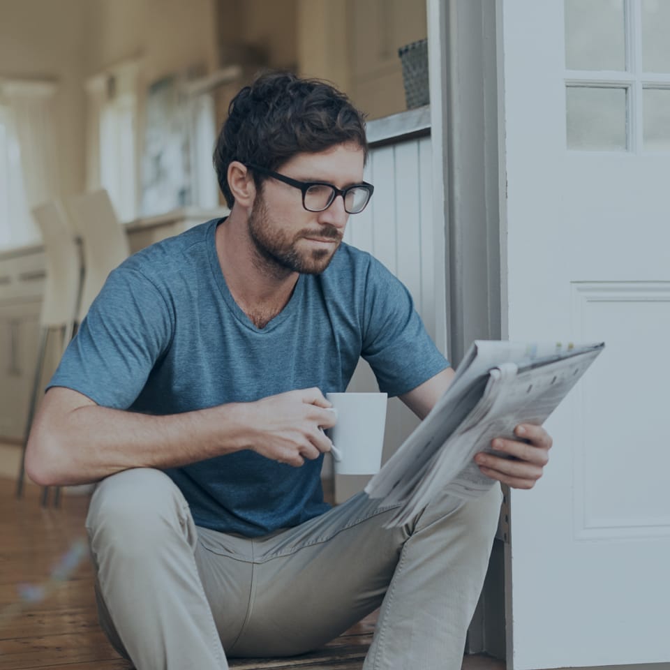 A man wearing glasses sits on the floor, holding a white mug in one hand and reading a newspaper with the other. He is dressed casually in a blue t-shirt and beige pants. The setting appears to be a bright, modern kitchen with wooden floors and white furniture.
