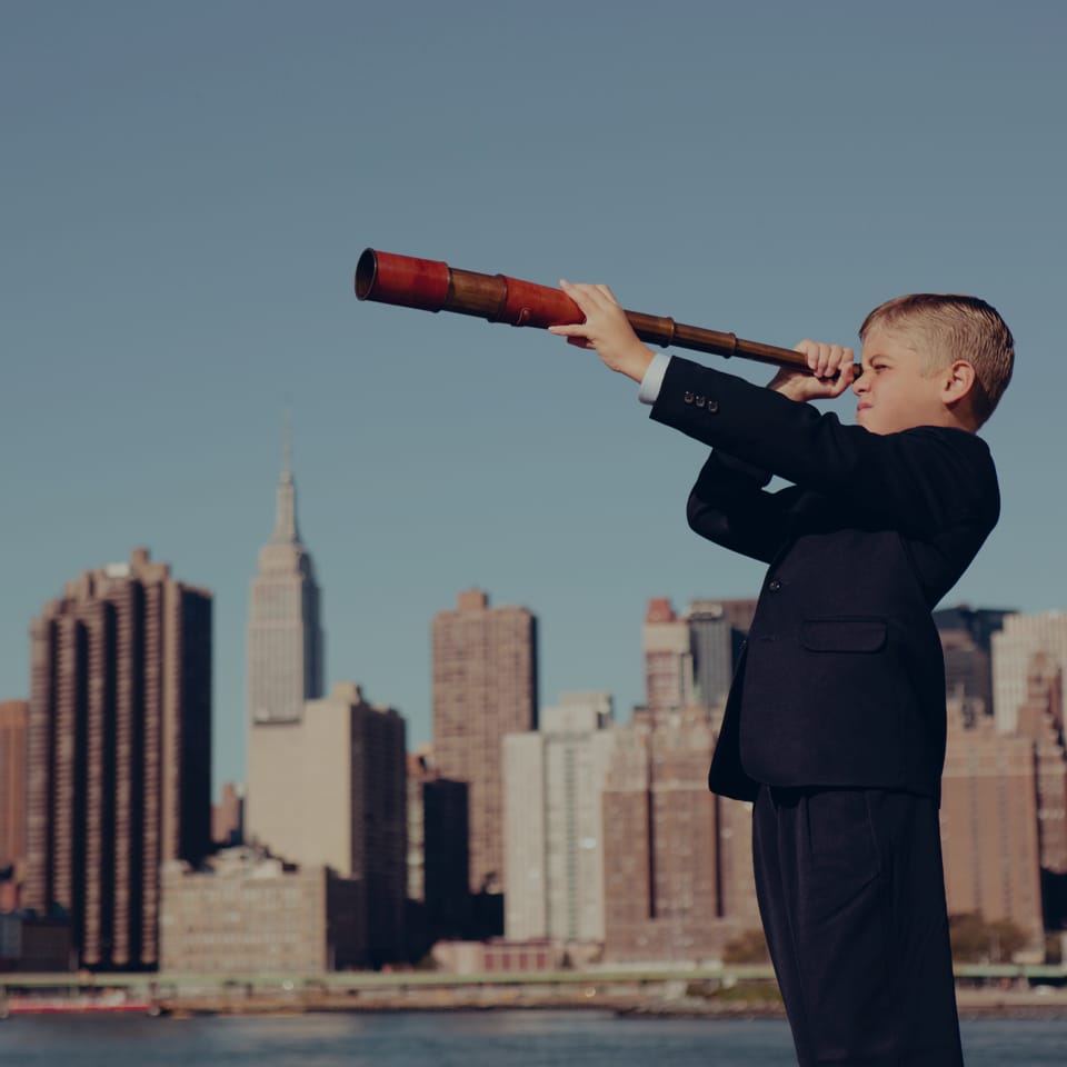 A young boy in a suit stands outdoors on a sunny day, holding and looking through an old-fashioned wooden telescope. The skyline of a city with tall buildings is visible in the background under a clear blue sky.