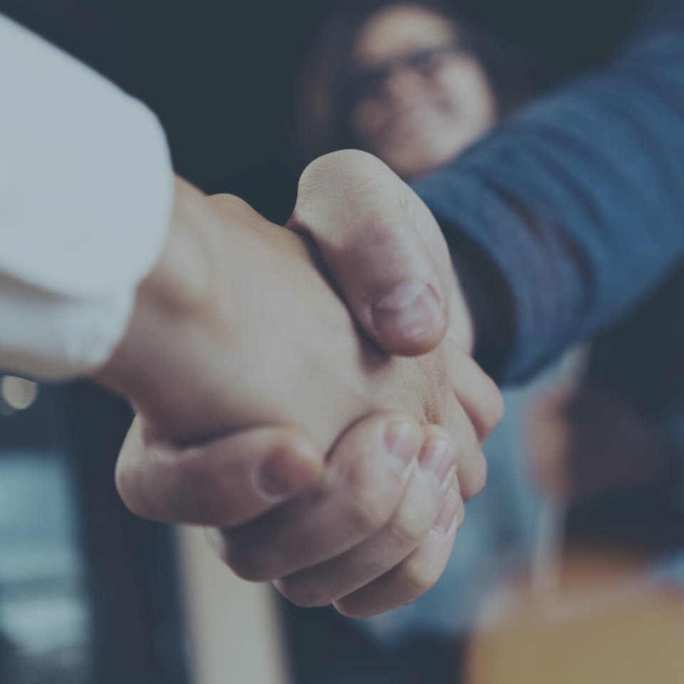 Close-up image of two people shaking hands, one wearing a white shirt and the other a blue long-sleeved garment. A blurred background suggests an indoor setting, with a third person slightly visible and out of focus.