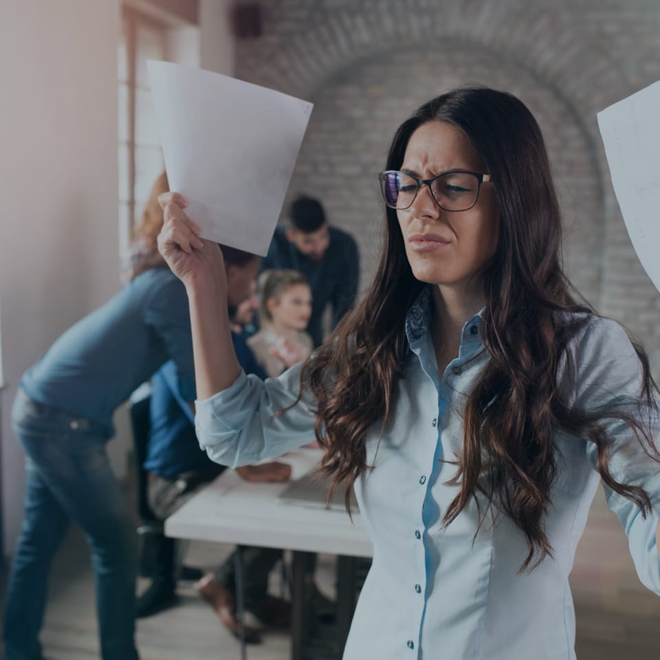 A frustrated woman wearing glasses and a light blue shirt holds up two sheets of paper in each hand. In the background, a group of people is gathered around a table, seemingly in a discussion in a room with brick walls and large windows.