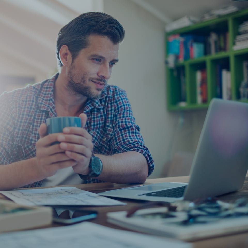 A man with short hair and a beard is sitting at a desk, holding a striped coffee mug and looking at a laptop screen. He is wearing a plaid shirt and a wristwatch. The desk has several papers and a pair of eyeglasses on it. A green shelf with books is in the background.