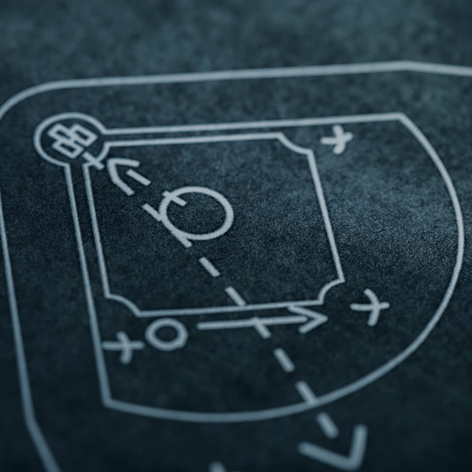 A close-up of a sports strategy chalkboard features a diagram with arrows, lines, and symbols. The drawing appears to be a play or tactic map, possibly for a ball game. The board has a worn texture and is primarily dark in color.