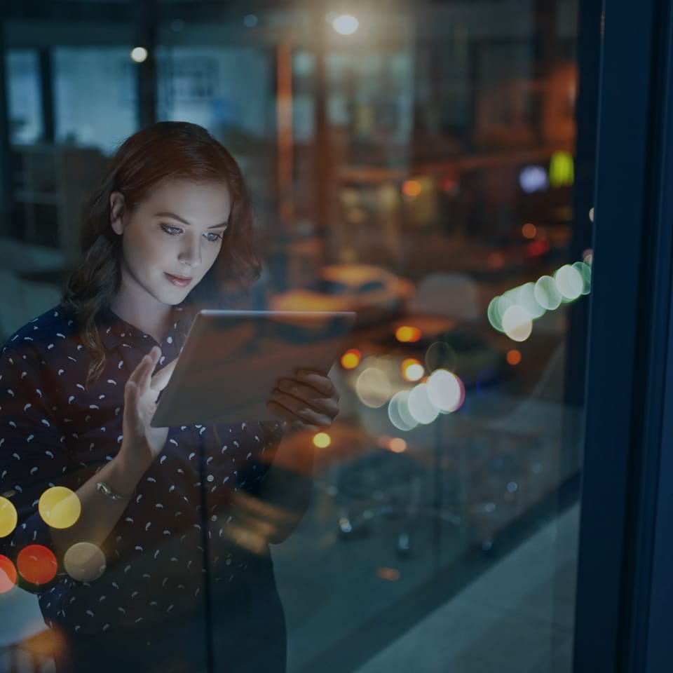 A woman standing by a window at night, holding and looking at a tablet. Outside, city lights create a bokeh effect through the glass, suggesting an urban environment. She appears focused on the screen, with reflections of the city adding depth to the scene.