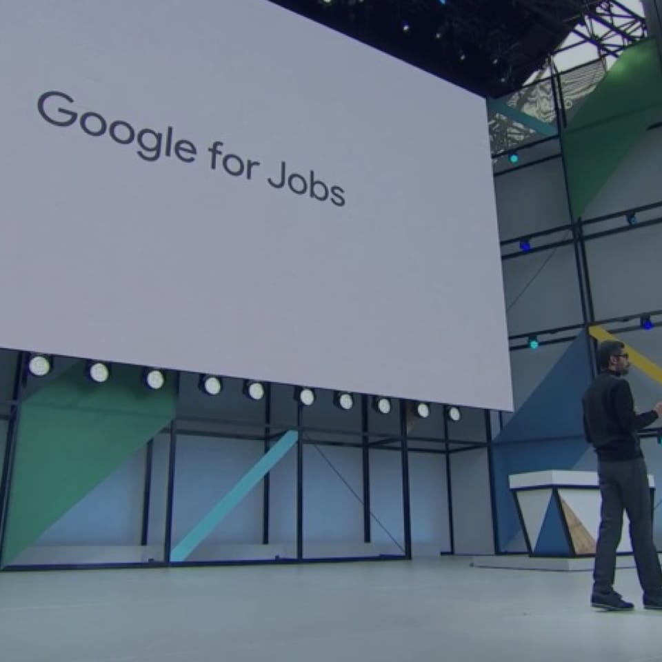 An individual is speaking on stage next to a large screen displaying the words "Google for Jobs." The stage setup is modern with geometric patterns and professional lighting.