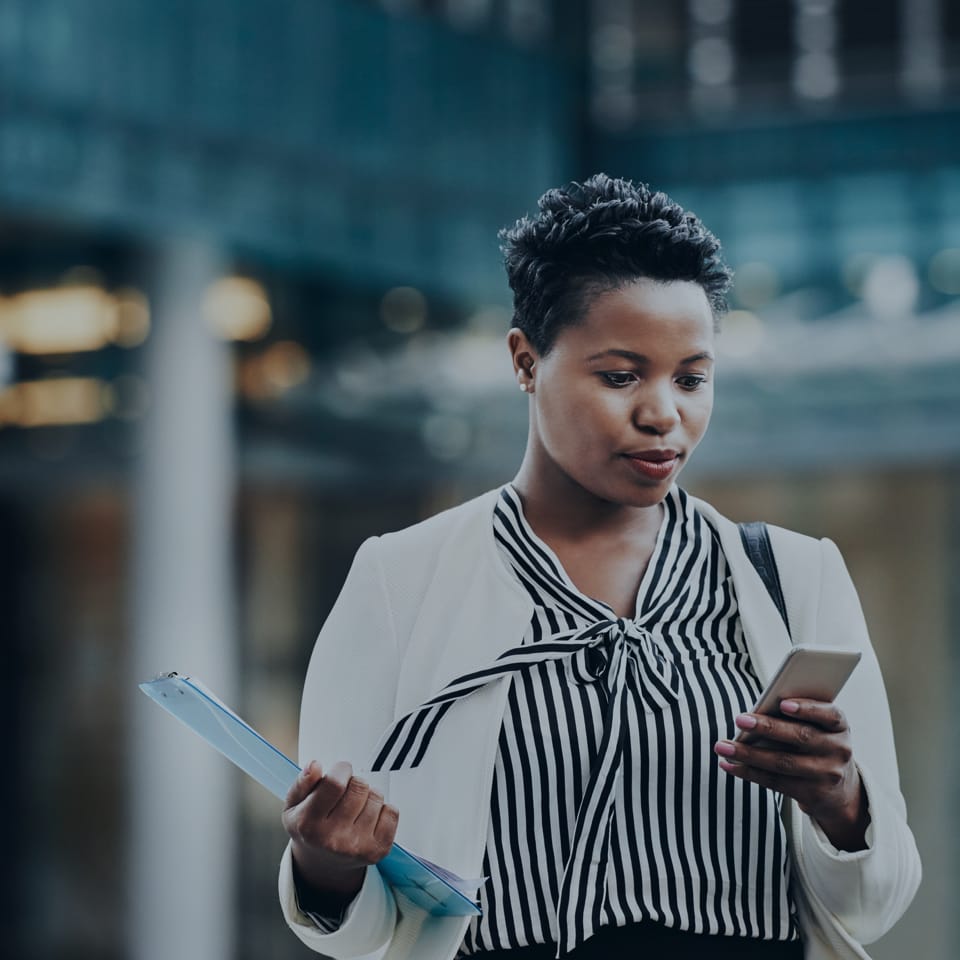 A woman wearing a striped blouse and white blazer is walking while looking at her smartphone. She is holding a blue clipboard in her left hand and a black shoulder bag is visible. The background is blurred, showing a modern building.