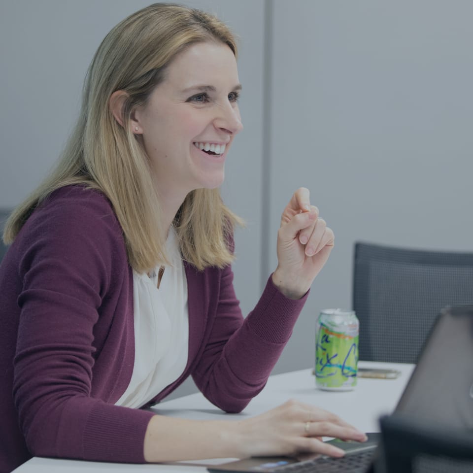A woman with long blond hair, wearing a white blouse and a maroon cardigan, smiles while seated at a desk. She has a can of flavored water near her and is using a laptop.
