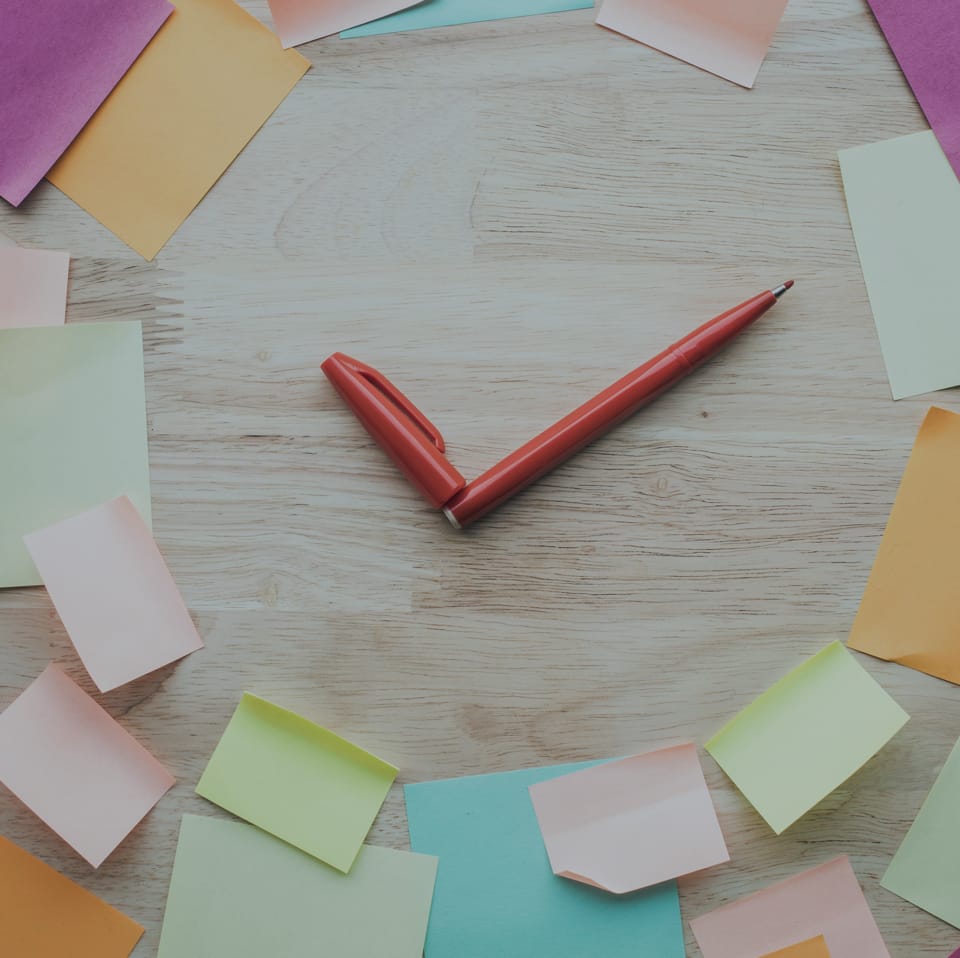 A red pen lays diagonally on a light wooden surface, surrounded by colorful sticky notes in shades of pink, orange, yellow, green, and blue, arranged in a loose circle around it.