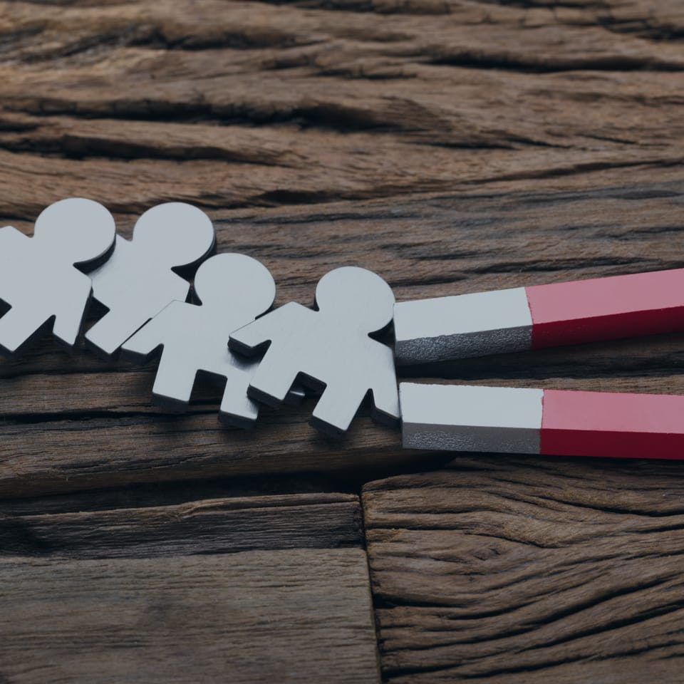 A U-shaped red and gray magnet attracts four small, white cut-out figures of people, pulling them across a textured wooden surface. The figures are aligned in a row, demonstrating the magnetic force in action.