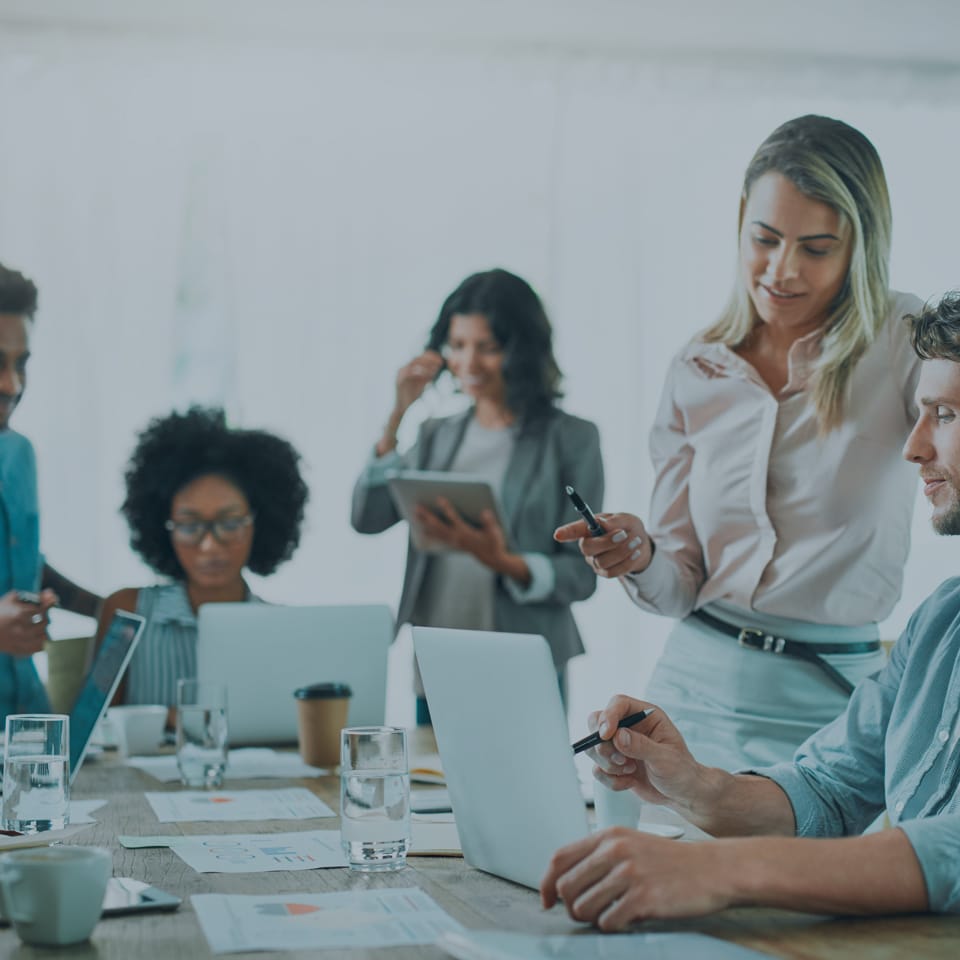 A group of people are gathered around a table in an office, each focused on their laptops and documents. One person is standing, pointing at a screen, while others are seated working. The atmosphere is collaborative and professional.