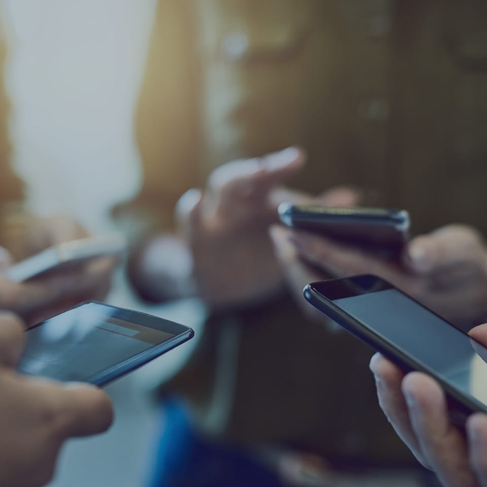 A group of people holding and using smartphones, focusing on their hands. The background is slightly blurred, emphasizing the devices and interaction, suggesting a collaborative or social activity.