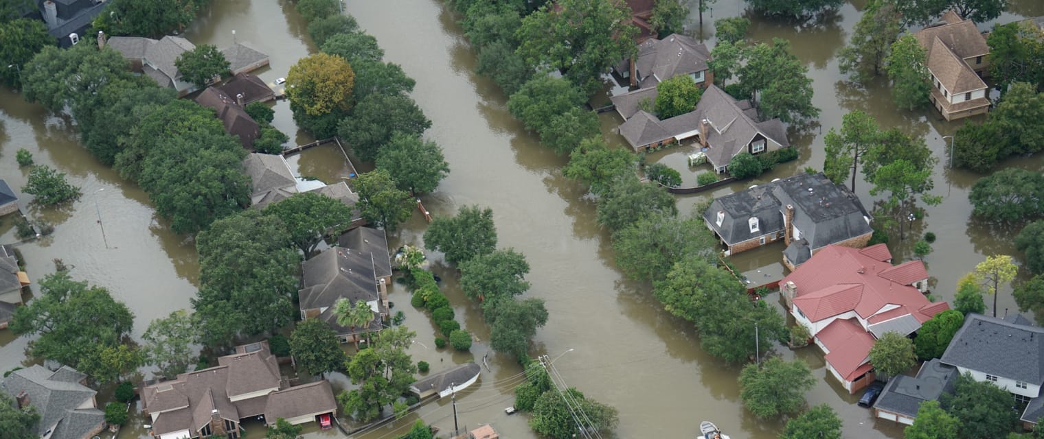 An aerial view of a flooded residential area with numerous houses surrounded by water. Streets are submerged, and trees are partially underwater. The scene captures the widespread impact of the flooding on the neighborhood.