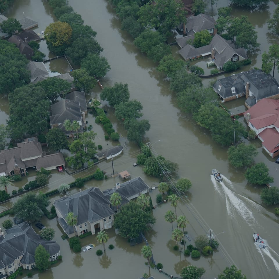 Aerial view of a flooded residential area with houses partly submerged in water. Streets are filled with water, and rescue boats navigate through the neighborhood. Trees and power lines are visible among the flooded homes.
