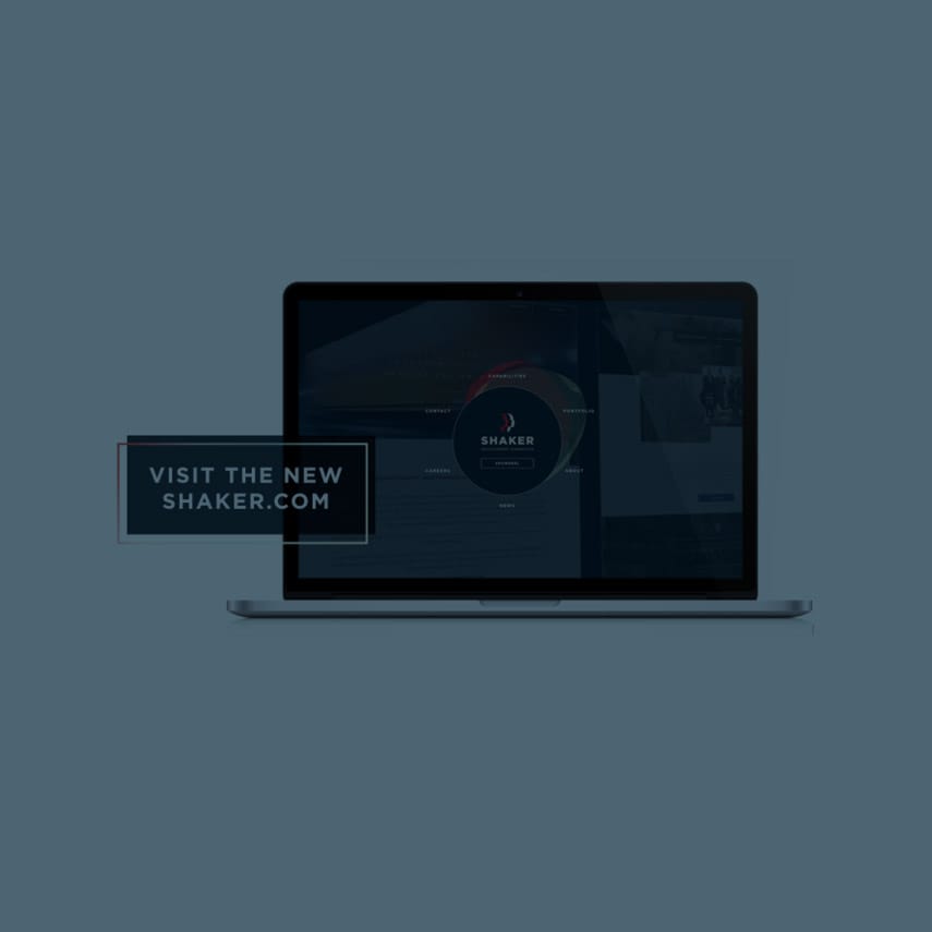A dark-themed image features an open laptop displaying a website with a circular logo in the center of the screen. A small rectangular pop-up box in the foreground contains the text: "VISIT THE NEW SHAKER.COM." The background is blurred out.