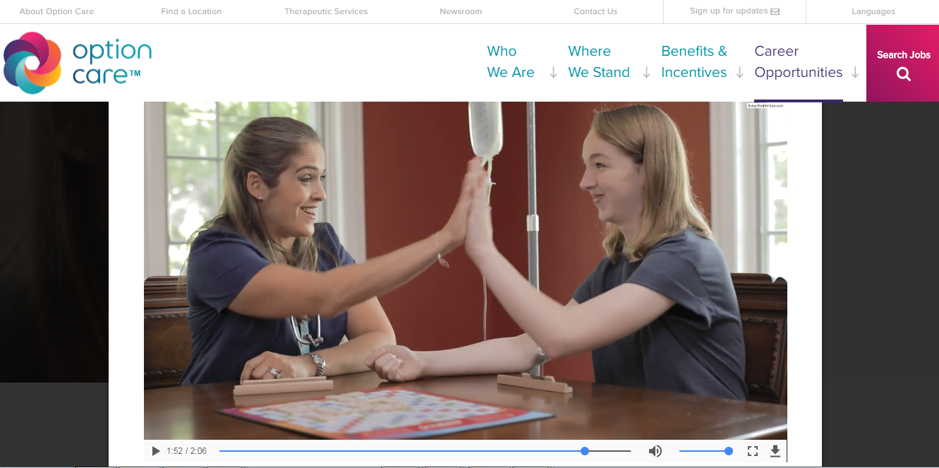Two people sit at a table playing a board game and high-fiving each other. Medical equipment, including an IV stand, is visible beside the table. The setting appears to be a home or clinic. A website's navigation menu and video control bar are also present on the screen.