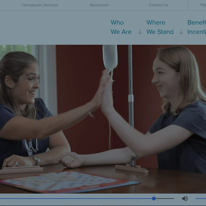 Two women, both wearing medical uniforms, sit at a table and high-five while smiling. One of them has an IV drip attached, indicating a medical setting. The background shows part of a webpage with text and navigation links.