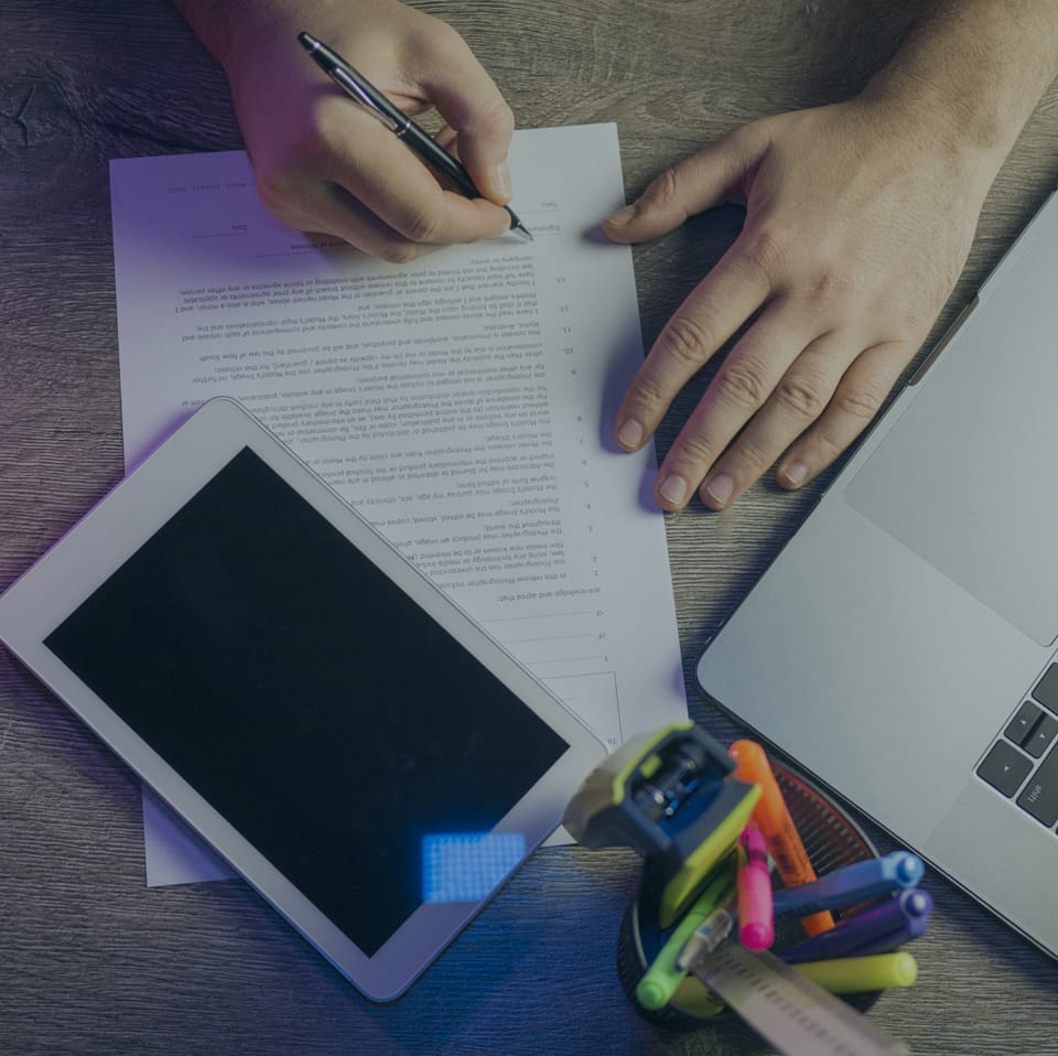 A person is writing on a document placed on a wooden desk. A tablet, a laptop, and a container holding pens, markers, and a ruler are also on the desk. One hand holds a pen, while the other rests on the document.
