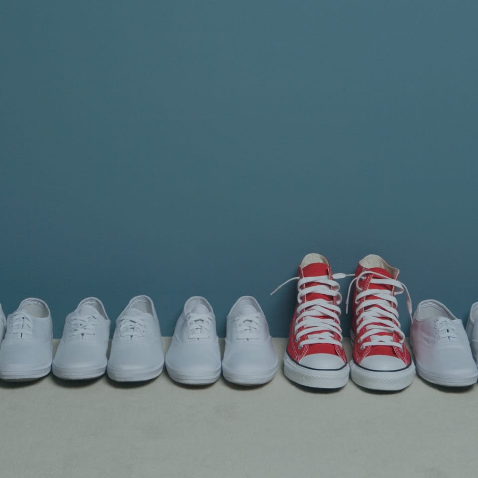 A row of white sneakers is lined up against a blue background, with a single pair of red high-top sneakers standing out among them. The image highlights the contrast between the uniform white shoes and the distinctive red ones.