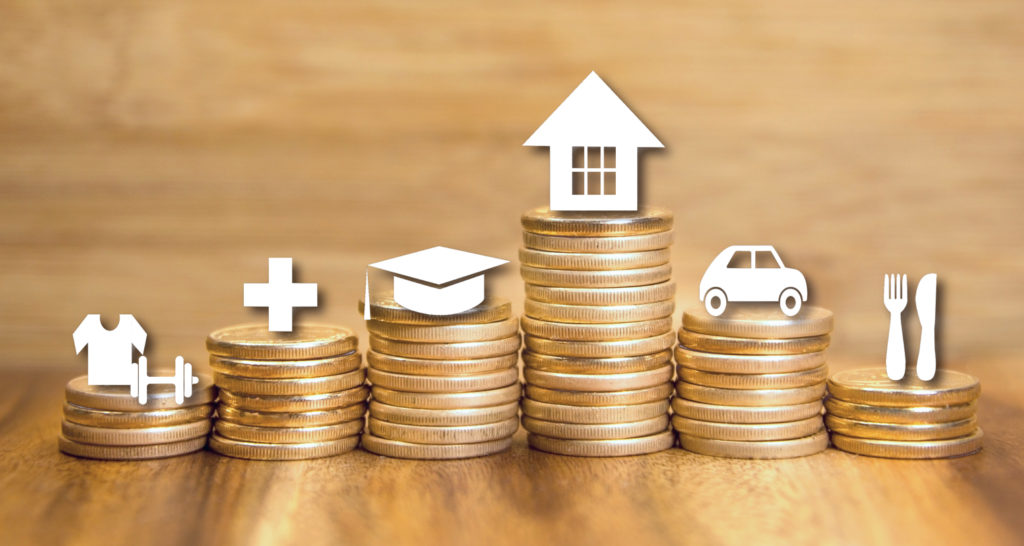 A row of ascending stacks of coins with white icons above each stack. From left to right, the icons depict clothing, healthcare, education, housing, a car, and dining, symbolizing various expenses and savings goals. The background is a warm, wooden texture.