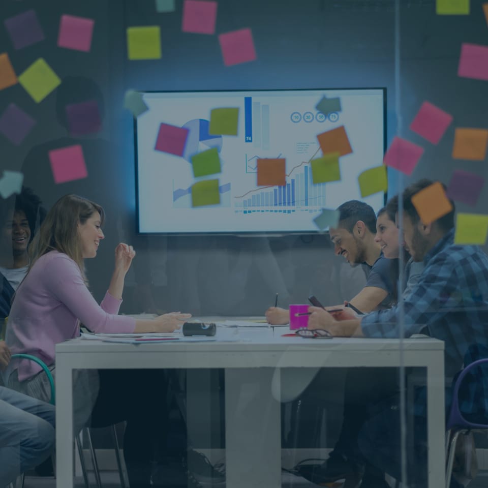 A group of five people sit around a table in a modern office, engaged in a meeting. Colorful sticky notes cover a glass wall. A large screen displays charts and graphs in the background. The atmosphere is collaborative and lively.