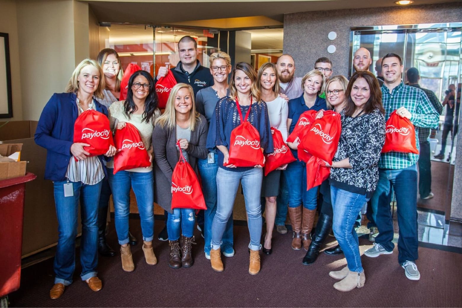 A group of 15 people is standing indoors, posing and smiling for a photo. They are holding red drawstring bags with the word "Paylocity" printed on them. The group appears cheerful and is casually dressed. The setting seems to be a corporate or office environment.