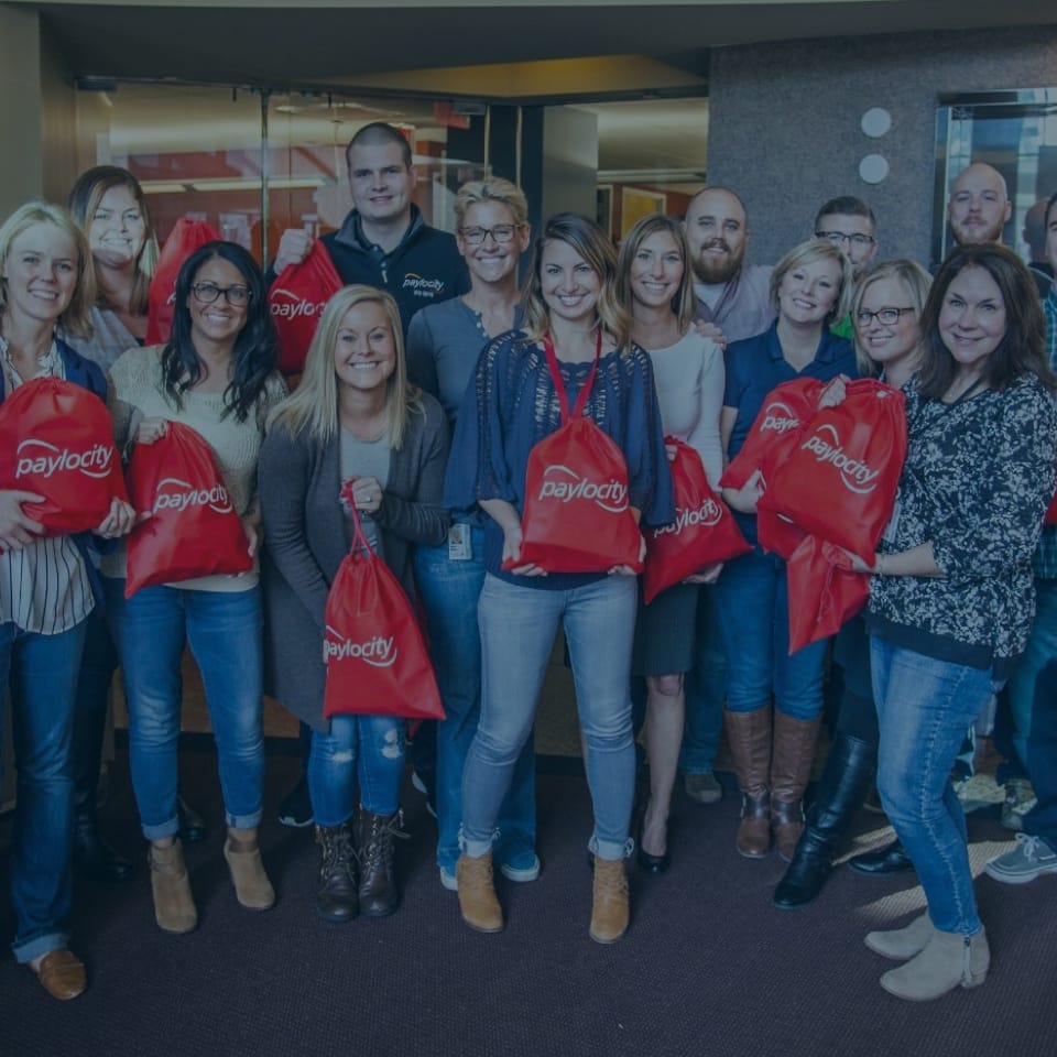 A group of smiling people is standing together indoors, holding red bags labeled "Paylocity." They appear to be in an office setting. Some people are wearing casual clothing, while others are dressed more formally.