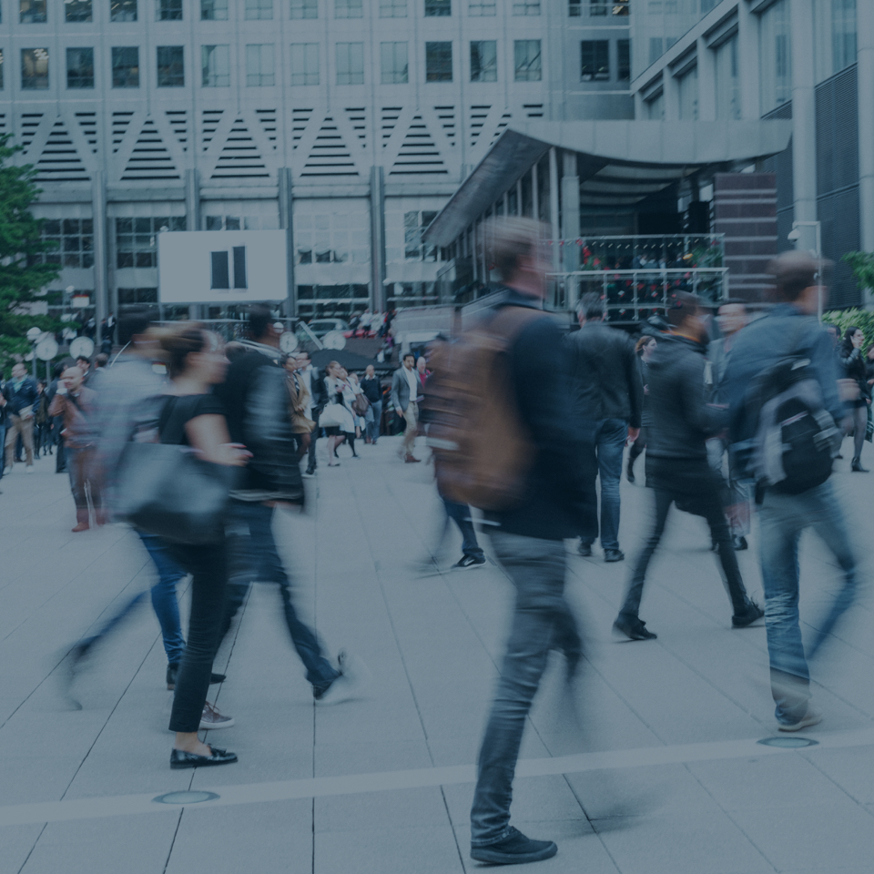 A bustling city square with numerous people walking in different directions. The buildings in the background suggest a modern urban environment. People are dressed in everyday attire, carrying backpacks and bags, showcasing the daily rush of urban life.