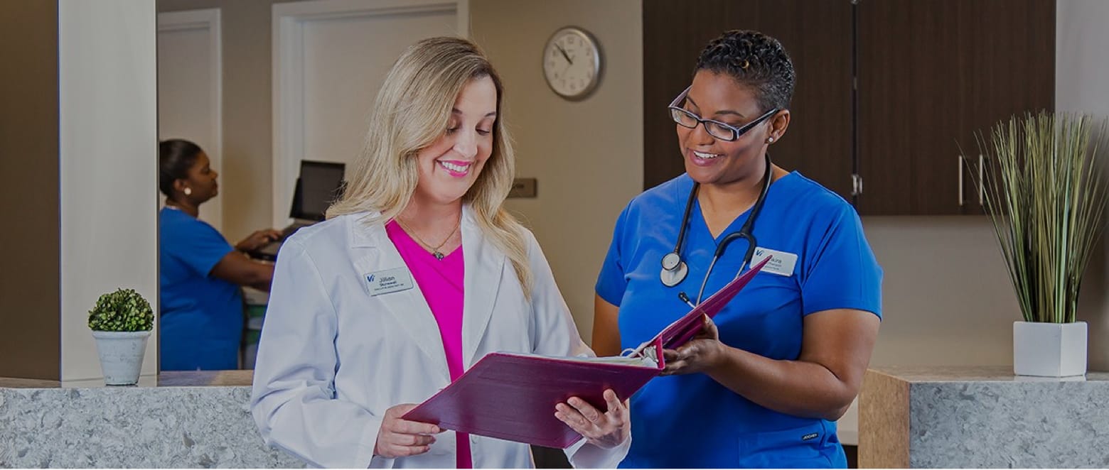 Two healthcare professionals are standing near a reception desk. One is wearing a white coat and holding a burgundy folder, smiling while the other, dressed in blue scrubs and a stethoscope, looks on. A potted plant and another professional are visible in the background.