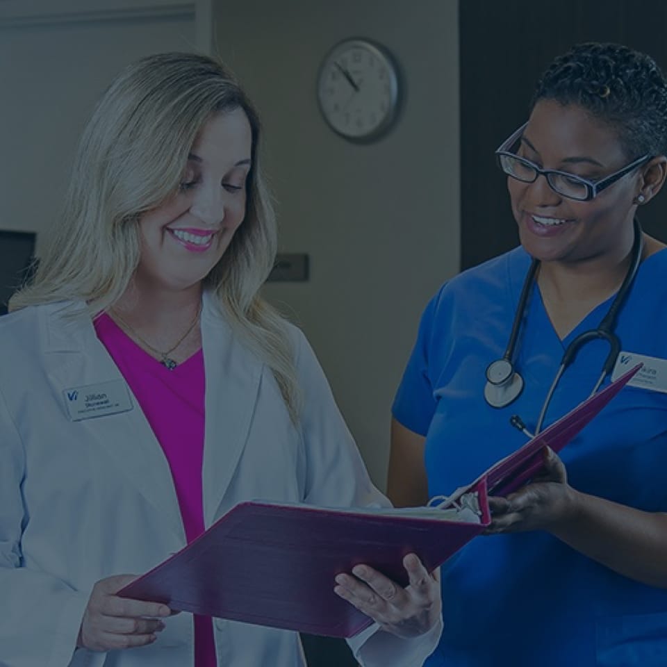 Two medical professionals, one in a white coat and the other in blue scrubs, smile while reviewing a red folder. They stand in a well-lit room with a clock on the wall behind them, indicating a collaborative and positive work environment.