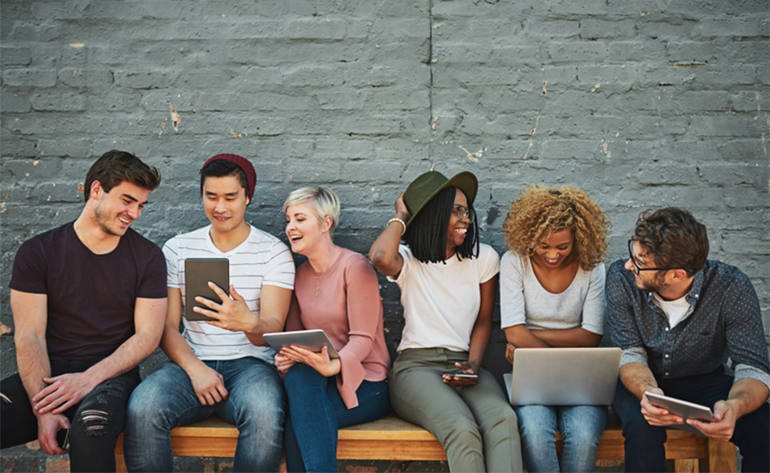 A diverse group of six friends sits on a wooden bench against a brick wall, laughing and engaged with digital devices such as smartphones, tablets, and a laptop. They appear relaxed and cheerful, with some looking at their screens and others interacting.