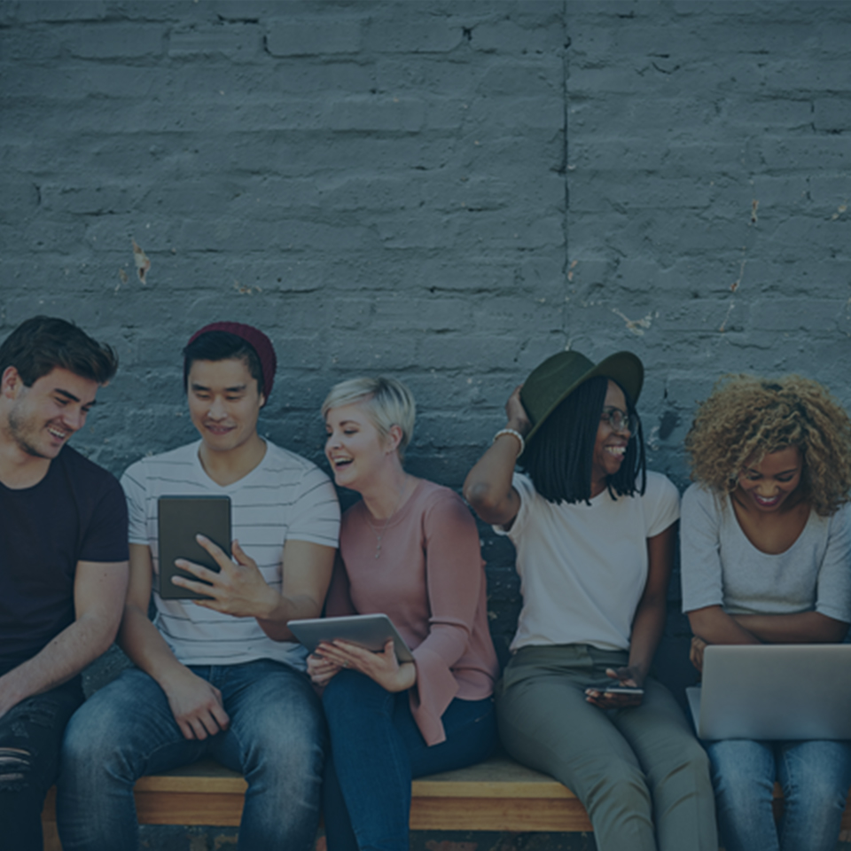 A diverse group of five young adults is sitting on a bench against a brick wall, casually interacting with each other. Each person is engaged with a different electronic device, such as a phone, tablet, and laptop. They appear to be enjoying their time together.
