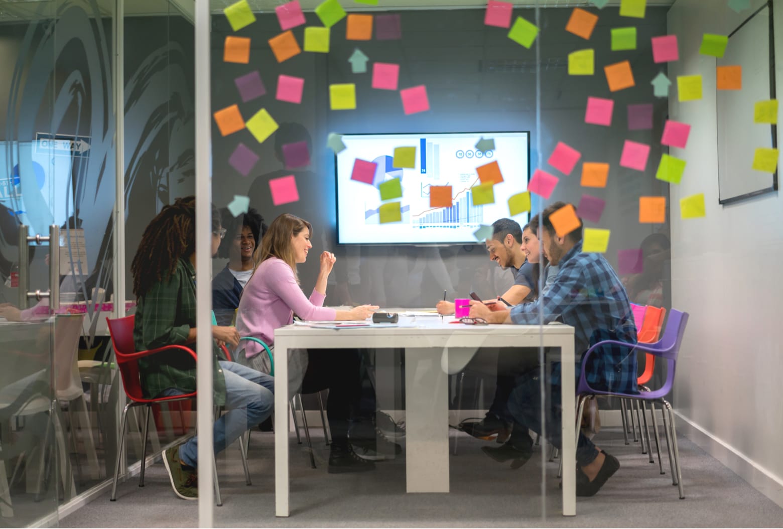 A group of people are sitting around a table in a glass-walled meeting room, engaged in discussion. A large screen displaying graphs and charts is visible on the wall. The glass walls are covered with colorful sticky notes.