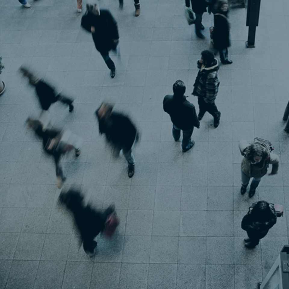 An overhead view of people walking in a public space with a tiled floor. Some people are standing still, while others appear blurred due to motion. The scene has an overall busy and dynamic atmosphere.