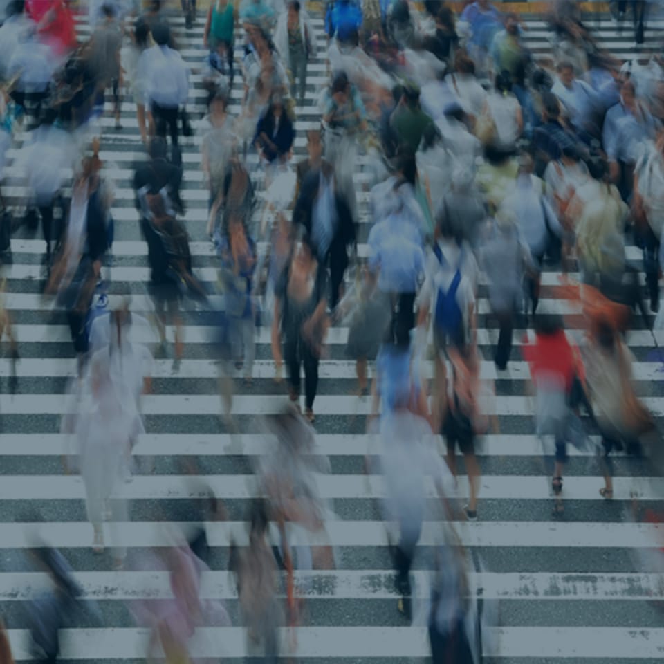 A bustling crowd of people hurriedly crossing a wide, striped crosswalk in an urban setting. The image is slightly blurred, conveying a sense of movement and urgency as individuals go about their day. Mixed colors and varied outfits indicate a diverse group.