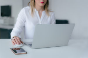 A person with light-colored hair wearing a white blouse is sitting at a table, using a laptop to browse Glassdoor. A smartphone rests on the table next to their hand, which is holding a computer mouse. The background is blurred, giving the impression of a modern workspace.