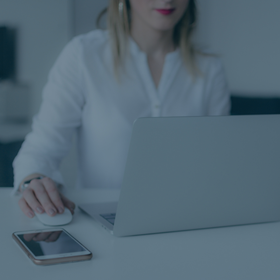 A person in a white shirt is using a laptop with one hand on a computer mouse. A smartphone is placed on the table next to the laptop. The background appears to be an office or work environment, reminiscent of a Glassdoor review scene. The image has a subtle, cool-tone filter.