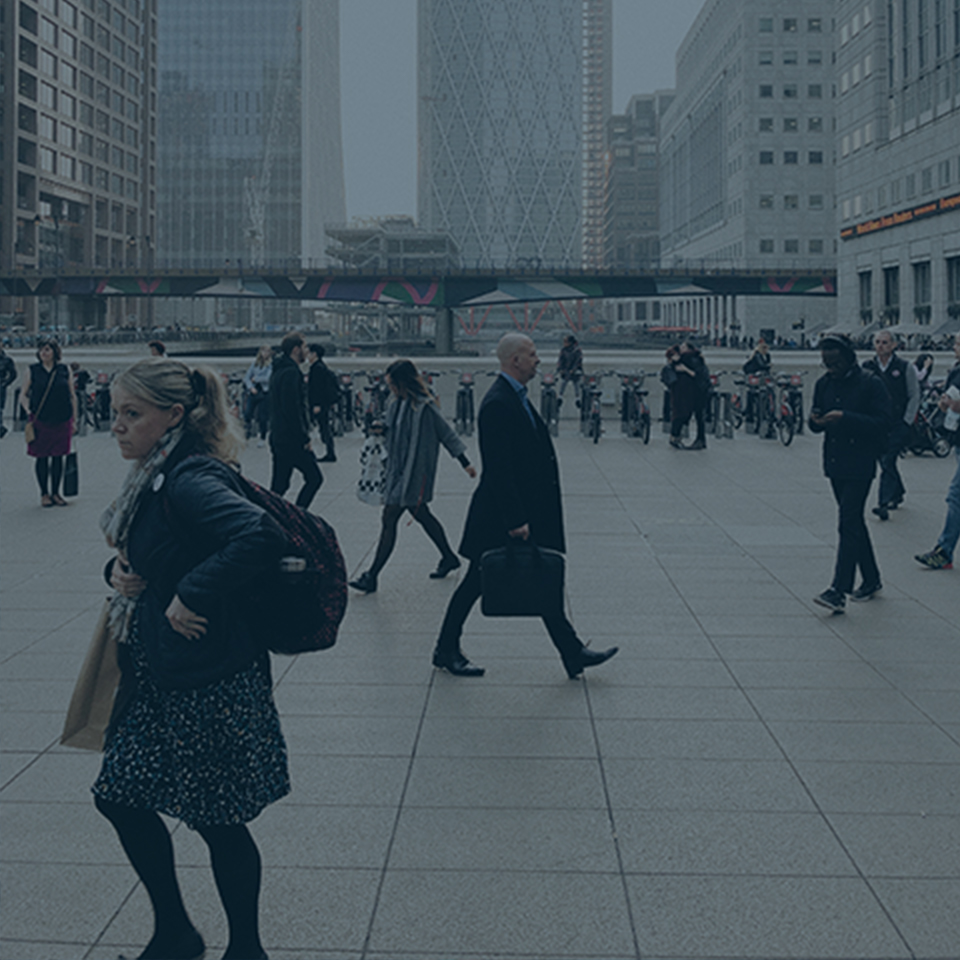 A busy urban square featuring people walking in various directions. In the background, modern glass buildings and an elevated walkway are visible. The pedestrians, dressed mostly in business attire, reflect the bustling job market on a typical workday in the city.