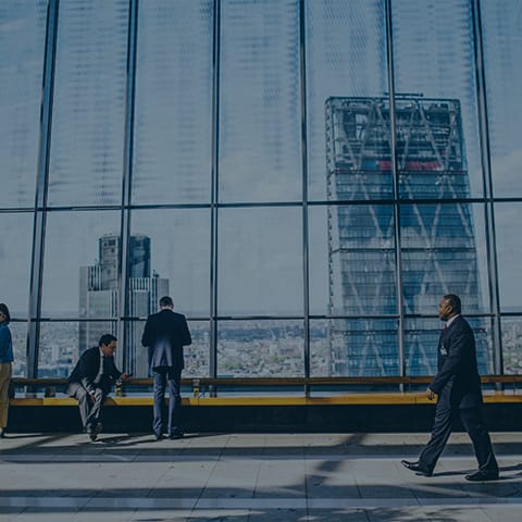 A large, modern office with floor-to-ceiling windows showcasing a cityscape. Several people in business attire are present: one stands looking out the window, another sits on a bench, and a third walks by. Skyscrapers are visible outside the windows.