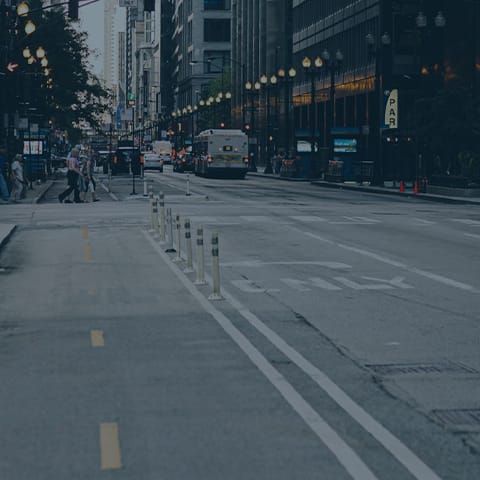 A city street scene showing a nearly empty road with a few cars in the distance. The street is lined with tall buildings, and there are streetlights along the sidewalk. A couple of pedestrians are visible crossing the street.