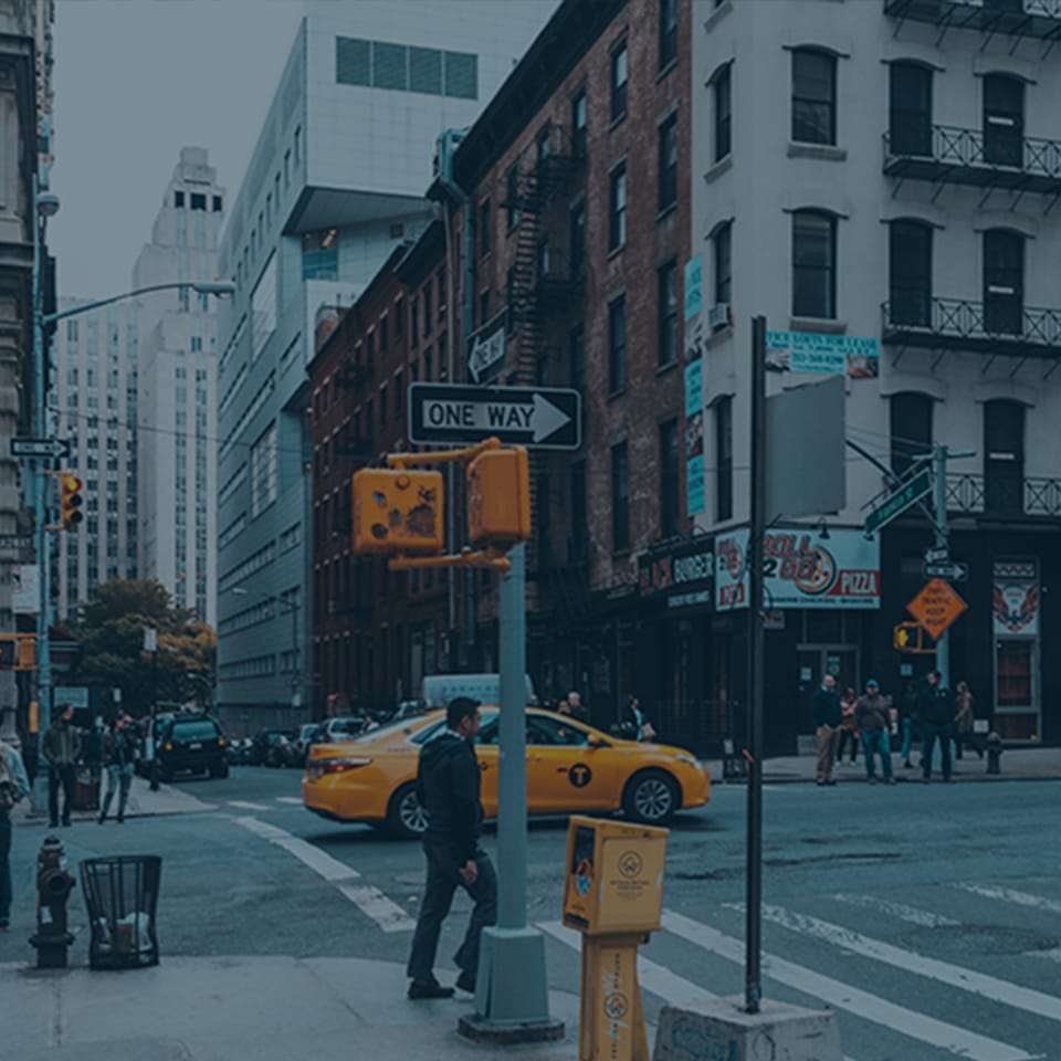 A busy city street corner showing a yellow taxi cab driving through the intersection. Pedestrians cross the street, and tall buildings, including a mix of modern and older brick structures, surround the area. Traffic signs and signal lights are visible.