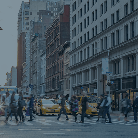 A busy city street with several people walking across the pedestrian crossing. Multiple yellow taxis are visible on the road, and tall buildings line both sides of the street. The sky is overcast but the scene is well-lit.