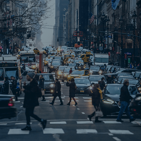 A busy city street filled with dense traffic, including cars, buses, and taxis. Pedestrians are crossing the road in various directions. Buildings and streetlights line the street, and bare tree branches are visible amidst the urban setting.