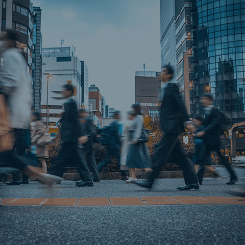 A busy urban street with several people in business attire walking briskly in different directions. Tall buildings and city structures are visible in the background under a cloudy sky, suggesting a typical workday in a city. The image has a slightly blurred effect.