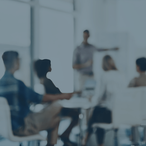 A group of five people sit around a table in a modern office setting with large windows. They are in a meeting or discussion. One person is standing near a whiteboard, gesturing, and appears to be speaking to the seated individuals. The image is blurred.