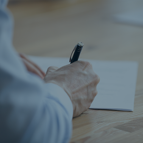 A close-up of a person’s hand holding a pen, writing on a piece of paper resting on a wooden table. The individual is wearing a light blue shirt with the sleeve slightly rolled up. The background is blurred.