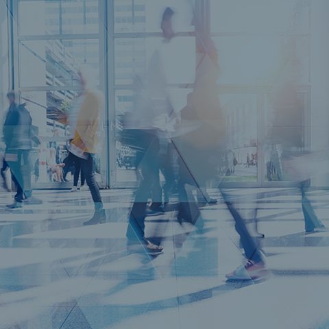 A blurred image of people walking through a spacious, brightly lit indoor area with large glass windows. The scene suggests a busy, bustling environment, possibly a modern office building or a commercial space. Light reflections and streaks add a dynamic effect.