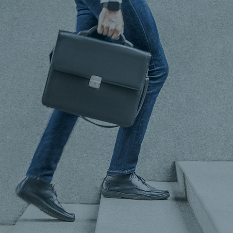 A person wearing blue jeans, black shoes, and a wristwatch is climbing gray stairs while carrying a black leather briefcase. The image focuses on the lower half of the person, highlighting the briefcase and their attire.
