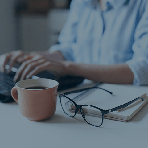 A person in a striped shirt is typing on a laptop keyboard. In the foreground, there is a pink coffee mug, a pair of black-rimmed glasses, and a spiral-bound notebook on the desk. The image has a soft, out-of-focus quality.
